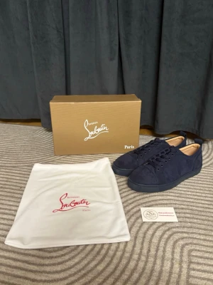 Mörkblå sneakers från Christian Louboutin - Snygga mörkblå sneakers från Christian Louboutin i mocka med klassisk röd sula och ton-i-ton snörning. Skorna har en stilren och minimalistisk design med diskreta sömmar och rundad tå. Perfekta för dig som vill ha något exklusivt och trendigt.