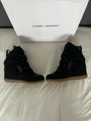 Isabel Marant skor  - Svarta sneakers från Isabel Marant med högt skaft och kilklack. Skorna har en cool design med kardborreband och detaljer i mocka och läder. Yttersulan är i gummi med beige ton. Perfekta för dig som vill ha en trendig och edgy look.
