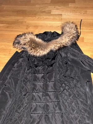 Svart parkas med äkta päls! - Snygg svart parkas med quiltad insida och stor huva med fluffig äkta päls i bruna och beige toner. Jackan har dragkedja och flera fickor, perfekt för kalla dagar. Klassisk modell som håller dig varm och stilren hela vintern. Jackan är köpt på Rocco Baroco men bytte ut fusk pälsen mot en riktig päls! 