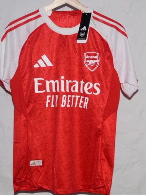 Arsenal röd matchtröja från Adidas - Snygg röd Arsenal matchtröja från Adidas med vita detaljer på axlar och ärmar. Tröjan har klubbmärke och Adidas-logga på bröstet samt 'Emirates Fly Better' tryckt framtill. Tillverkad i lätt och ventilerande material, perfekt för fotbollsträning. Är du efter en annan storlek är det bara att skriva!
