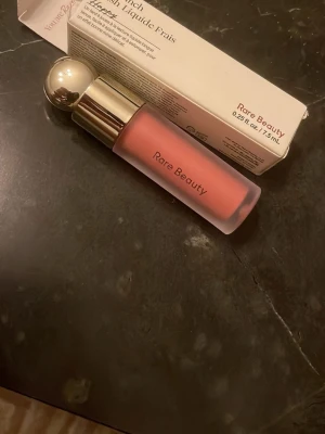 Rare Beauty Soft Pinch Liquid Blush - Flytande rouge från Rare Beauty i nyansen Soft Pinch. Kommer i en frostad glasflaska med guldfärgat lock och praktisk applikator. Färgen är en varm, persikorosa ton som ger ett naturligt fräscht glow till kinderna. Perfekt för dig som gillar ett mjukt och lätt resultat.