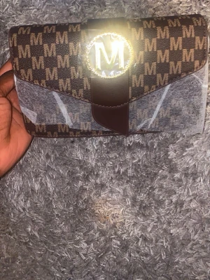 Brun kuvertväska med M-mönster - Snygg brun kuvertväska med guldfärgad M-detalj framtill och monogrammönster över hela väskan. Väskan har magnetlås, innerfack med dragkedja och medföljande axelrem i läderimitation. Perfekt för dig som gillar trendiga accessoarer.