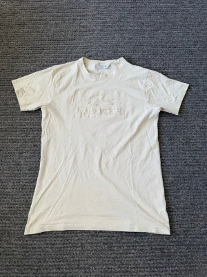 T-shirt - Snygg beige t-shirt från Lacoste med klassiskt relieftryck av krokodilen och loggan på bröstet. T-shirten har rund halsringning och korta ärmar. Tillverkad i mjuk bomull för en skön känsla och clean look.