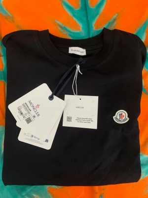 Svart Moncler t-shirt med logga - Snygg svart t-shirt från Moncler med klassisk rund hals och Moncler-logga broderad på bröstet. Tillverkad i mjuk bomull som känns skön mot huden. Perfekt för dig som vill ha en stilren och enkel look.