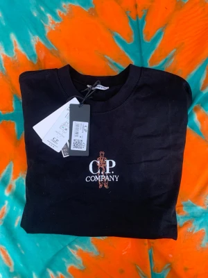 Svart C.P. Company t-shirt - Snygg svart t-shirt från C.P. Company med stor logga och grafiskt tryck på bröstet och ryggen. Klassisk rund hals och korta ärmar. Perfekt för dig som gillar streetwear och vill ha något stilrent men ändå med edge.