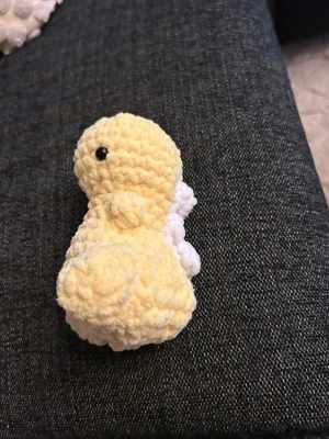 Gul virkad dinosauriefigur - Söt handvirkad dinosaurie i mjukt gult garn med små vita vingar på ryggen och svarta ögon. Perfekt som dekoration eller leksak. Figuren har rundad form och är stoppad för att vara mjuk och gosig.