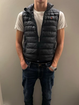 Svart dunväst från Polo Ralph Lauren - Snygg mörkblå dunväst från Polo Ralph Lauren med klassisk röd logga på bröstet. Västen har dragkedja framtill, två fickor med dragkedja och en hög krage. Perfekt att slänga över en hoodie eller t-shirt. Storlek L i barn (14-16)år den är dock lite liten i storleken. Samt att mitt namn står i den.