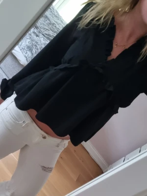 Svart blus med volangdetaljer - Snygg svart blus med volangdetaljer längs ringningen och framtill. Blusen har en lös passform och långa ärmar. 
