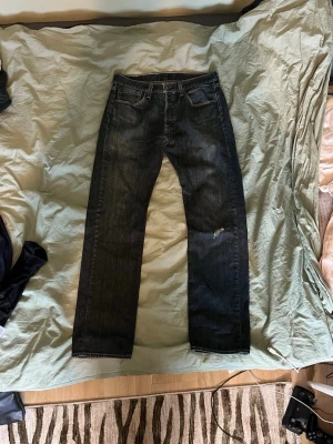 Mörkblå 501 Levi's jeans - Säljer ett par mörkblå 501 jeans från Levi's med klassisk femficksdesign och en liten slitning på ena benet. Jeansen har en tydlig läderpatch bak i midjan och är tillverkade i kraftig denim. Perfekta för en avslappnad och tidlös look. Storlek 31/32. Hör av dig vid minsta fundering!