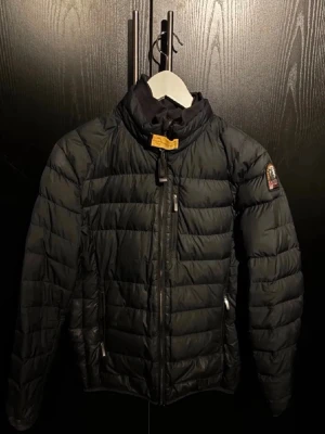 Svart dunjacka från Parajumpers - Snygg svart dunjacka från Parajumpers med klassisk patch på ärmen. Jackan har dragkedja framtill, hög krage och två sidofickor med dragkedja. Perfekt för kalla vinterdagar tack vare det varma dunfodret.