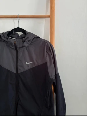 Nike svart och grå vindjacka - Snygg vindjacka från Nike i svart och mörkgrått med dragkedja framtill och huva. Jackan har två sidofickor och en reflekterande Nike-logga på bröstet. Perfekt för blåsiga dagar och sportiga outfits.