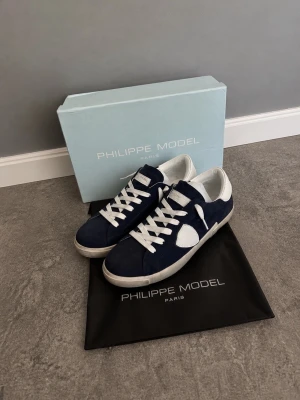 Philippe Model Low PRSX MÖRKBLÅ SUEDE  - Mörkblå sneakers från Philippe Model med vita detaljer och vita skosnören. Skorna har en klassisk låg siluett och är tillverkade i mocka och läder. Perfekta för en stilren och trendig look. Mörkblå skor från Philippe Model  modellen är mörkblå Low Prsx Suede, perfekt för hösten och vintern. 