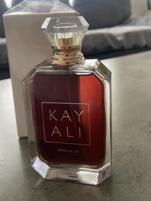 Kayali Vanilla 28 Eau de Parfum - Kayali Vanilla 28 Eau de Parfum i en elegant, genomskinlig glasflaska med guldiga detaljer och en kristalliknande kork. Doften har en djup bärnstensfärgad vätska och flaskan har en modern, lyxig design. Volymen är 100 ml.