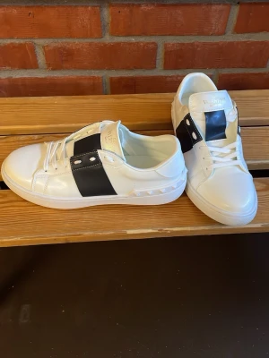 Vita sneakers med svart detalj Valentino - Snygga vita sneakers från Valentino med bred svart läderdetalj över sidan och nitar längs sulan. Klassisk låg modell med vita skosnören och guldtext på plösen. Perfekta för dig som vill sticka ut med stilrena detaljer.