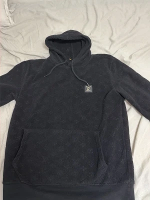Svart hoodie med monogram och huva - Mysig svart hoodie med huva och dragsnören, samt en stor magficka framtill. Hoodien har ett diskret monogrammönster över hela tyget och en patch med logga på bröstet. Perfekt för en chill och stilren look.