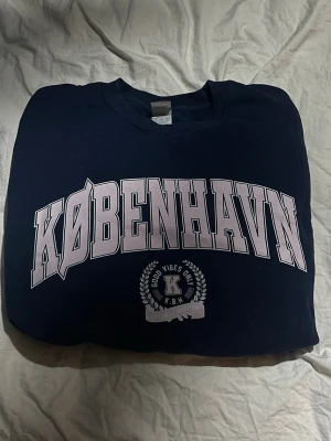 Mörkblå hoodie med KØBENHAVN-tryck - Snygg mörkblå hoodie med stort vitt KØBENHAVN-tryck på bröstet och ett mindre emblem med texten 'Good Vibes Only' och 'K.B.H' under. Klassisk passform och mjukt material, perfekt för en chill och trendig look.