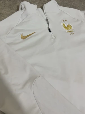 Vit Nike Frankrike träningströja - Snygg vit långärmad träningströja från Nike med Frankrikes landslagsemblem och två guldstjärnor på bröstet. Tröjan har halv dragkedja och guldfärgade detaljer, perfekt för fotbollsträning eller sportig stil.