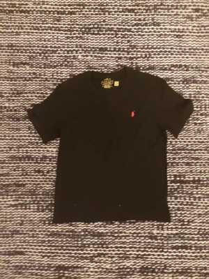 Svart t-shirt från Polo Ralph Lauren - Klassisk svart t-shirt från Polo Ralph Lauren med den ikoniska röda broderade loggan på bröstet. T-shirten har rund hals och korta ärmar, perfekt till alla tillfällen. Tillverkad i mjuk bomull för skön komfort.