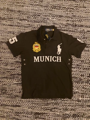Svart pikétröja med tryck från Polo Ralph Lauren - Svart pikétröja från Polo Ralph Lauren med vit logga och texten 'MUNICH' framtill. Tröjan har krage, knappar vid halsen och broderad detalj på bröstet samt siffran 5 på ärmen. Perfekt för en sportig och stilren look.