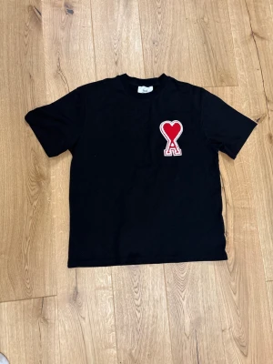 Svart t-shirt med rött hjärta från Ami - Svart t-shirt från Ami med ett stort rött hjärta och bokstaven A broderat på bröstet. Klassisk passform och rund halsringning. Perfekt för dig som gillar stilrena plagg med en twist.