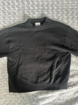 Svart stickad tröja med rund hals - En enkel och stilren svart stickad tröja med rund hals och ribbade muddar vid ärmslut och nederkant. Tröjan har en rak passform och är perfekt för lager på lager. Materialet ser mjukt och bekvämt ut.