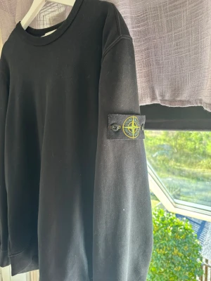 Svart sweatshirt från Stone Island - Säljer en svart sweatshirt från Stone Island med den klassiska logotypen på vänster ärm. Tröjan har rund halsringning, långa ärmar och ribbade muddar. Perfekt för dig som gillar streetwear och enkel stil. Bra skick och försiktigt använd. Står L i storlek men skulle säga L/XL