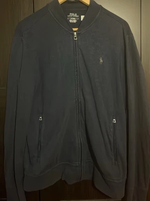 Mörkblå zip hoodie från Polo Ralph Lauren - Snygg mörkblå hoodie med dragkedja från Polo Ralph Lauren. Klassisk design med två fickor framtill och diskret logga på bröstet. Tillverkad i mjukt material och har ribbade muddar vid ärmslut och nederkant. Perfekt för en chill och stilren look.
