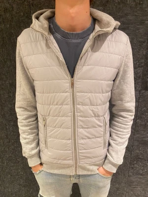 Ljusgrå hoodie från Reiss - Snygg ljusgrå hoodie med quiltad frontpanel och dragkedja. Ärmar och rygg i mjukt sweatshirtmaterial, medan framsidan har vadderad look. Två fickor med dragkedja och ribbade muddar. Perfekt för en chill och stilren vardagsoutfit.