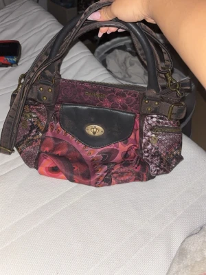 Mönstrad handväska från Desigual - Färgstark handväska från Desigual med unikt mönster i rosa, rött och lila toner. Väskan har detaljer i brunt konstläder, en svart ficka framtill med vridlås och dragkedjor i guld. Två handtag och en axelrem gör den enkel att bära.