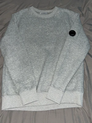 Grå sweatshirt från C.P. Company - Snygg grå sweatshirt från C.P. Company med rund halsringning och ribbade muddar. Tröjan har en ikonisk svart linsdetalj på ärmen som ger en cool streetwear-vibe. Perfekt till jeans eller cargo för en avslappnad look.