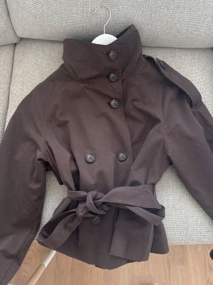 Mörkbrun trenchcoat med knytskärp bershka - Stilren mörkbrun trenchcoat med dubbelknäppning, hög krage och knytskärp i midjan. Jackan har axelklaffar och dekorativa knappar vid ärmsluten. Tillverkad i ett slätt tyg som ger en klassisk look. Perfekt för dig som vill ha en tidlös och trendig jacka.