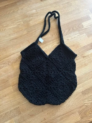 Svart virkad shoppingväska - Snygg svart shoppingväska i virkat material med unikt mönster och dubbla axelband. Väskan har en mjuk, flexibel form och är perfekt för dig som gillar en bohemisk och trendig stil. Rymmer mycket och är enkel att bära över axeln.