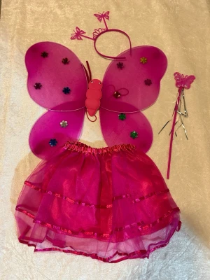 Fjäril maskeradset i 4 Delar  - Sött maskeradset med fjärilstema i rosa och lila. Innehåller vingar dekorerade med blommor, matchande kjol i tyll, diadem med fjärilar och trollstav. Perfekt för cosplay eller maskerad. Materialet är lätt och skimrande polyester. 145kr/set/bild , finns röd och blå färg kvar 