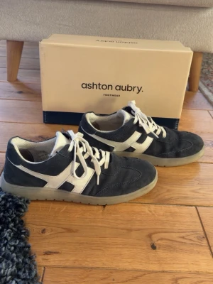 Mörkblåa och vita sneakers Ashton Aubry - Snygga sneakers från Ashton Aubry i mörkblå mocka med vita detaljer och vita skosnören. Skorna har en låg profil och genomskinlig sula. Knappt använda, och har låda kvar. Storlek 42. 