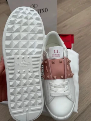 Valentino vita sneakers med nitar - Snygga vita sneakers från Valentino med ljusrosa band över vristen dekorerat med ikoniska nitar. Skorna har en chunky vit sula med rutmönster undertill och klassisk snörning. Tillverkade i läder för en lyxig känsla och stilren look.