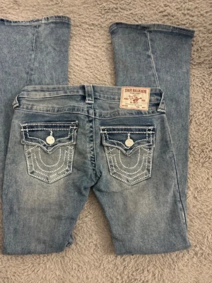 Blå True Religion jeans med detaljer - Säljer ett par blå jeans från True Religion med ikoniska fickor bak, dekorerade med vita sömmar. Perfekta för dig som gillar streetwear och vill sticka ut.
