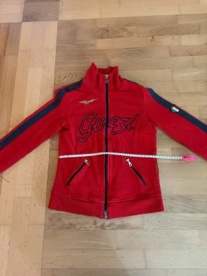 Röd Guzzi hoodie med dragkedja - Priset kan diskuteras😊Snygg röd hoodie med dragkedja framtill och svarta detaljer längs ärmarna. Stort Guzzi-tryck på bröstet, broderad logga och italiensk flagga på ena ärmen. Två fickor med dragkedja och hög krage. Perfekt för dig som gillar sportig stil. Tröjan har en liten lös tråd vid dragkedjan som inte påverkar användningen av tröjan.