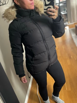 Svea jacka med fuskpäls - Snygg svart medellång jacka med dragkedja och stor go Luva. En innerficka, muddar i ärmar. Jackan har en hög krage och quiltad design som håller dig varm under kalla dagar. Dragsko längst ner så man kan reglera hur tajt man vill ha den. Perfekt för vinter och har en modern, avslappnad passform. St xs, men jag kan ha den , jag har st S. Enda fel är att dragkedjorna på fickorna fungerar ej. Men går att fixa. För övrigt har jackan inga fläckar, revor eller dylikt. Jättefin. 