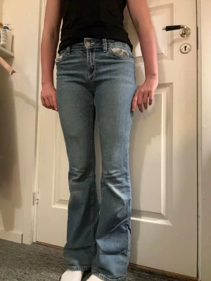 Ljusblå bootcut jeans - Säljer ett par ljusblå bootcut jeans med klassisk femficksdesign och snygga detaljer på bakfickorna. Jeansen har normal midja och är tillverkade i mjuk denim med lätt tvättad look. Perfekta för dig som gillar retrovibbar och avslappnad stil.