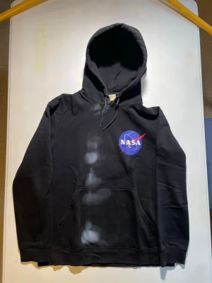 Svart NASA hoodie med tryck - Säljer en svart hoodie med NASA-logga tryckt både fram och bak. Hoodien har klassisk känguruficka och dragsko i huvan. Materialet ser ut att vara mjukt och bekvämt, perfekt för dig som gillar rymdtema och streetwear.
