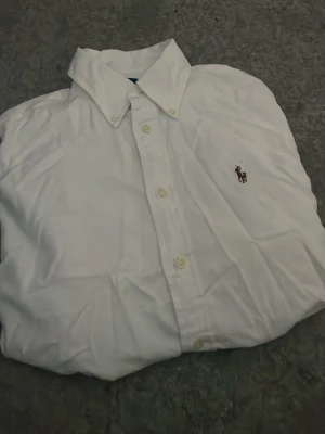 Vit skjorta från Ralph Lauren - Klassisk vit skjorta från Ralph Lauren med broderad logga på bröstet. Skjortan har knappar framtill och en tidlös design som passar till allt. Tillverkad i bomull för en skön och luftig känsla. Pris kan diskuteras vid snabb affär.