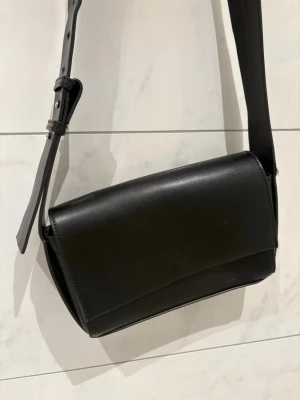 Svart stilren axelväska i läder NA-KD - Snygg svart axelväska i slätt läder med minimalistisk design och bred justerbar axelrem. Väskan har ett stilrent uttryck och passar perfekt till dig som gillar en clean look. Magnetstängning framtill och rektangulär form. Mycket bra skick, inga märken på utsidan🖤