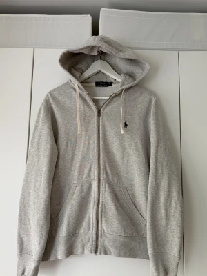 Grå zip hoodie från Polo Ralph Lauren - Snygg grå hoodie med dragkedja från Polo Ralph Lauren. Klassisk design med huva och vita dragsnören, samt broderad logga på bröstet. Mjuk bomullsblandning och två fickor framtill. Perfekt för en chill och stilren look.