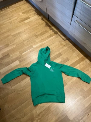Grön hoodie från Calvin Klein - Snygg grön hoodie från Calvin Klein med huva och lång ärm. CK-logga i vitt på bröstet ger en clean och modern look. Tillverkad i mjukt material som känns skönt mot huden. Perfekt för dig som vill ha en enkel men stilren hoodie.