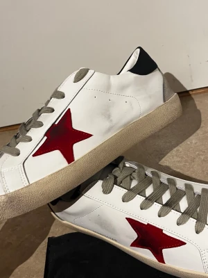 Golden Goose skor  - Säljer ett par vita sneakers med en stor röd stjärna på sidan, grå skosnören och beige sula. Skorna har svart detalj på hälen och texten GGDB/STAR på sidan. Materialet är läder och de har en klassisk låg modell.