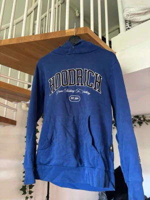 Blå hoodie från Hoodrich - Snygg blå hoodie från Hoodrich med stor logga och text på bröstet samt tryck på ryggen. Klassisk känguruficka framtill och huva. Perfekt för dig som gillar streetwear och vill sticka ut med en cool design.
