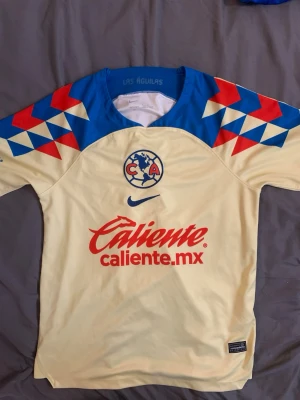 Club América matchtröja Nike gul/blå - Säljer en officiell matchtröja från Club América i gult med blå och röda detaljer på axlarna. Tröjan har klubbens logga på bröstet, Nike-logga och sponsortryck framtill. Tillverkad i lätt och ventilerande material, perfekt för fotboll eller träning.