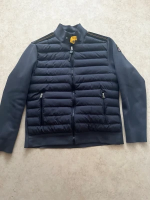 Mörkblå dunväst från Parajumpers - Snygg mörkblå dunväst från Parajumpers med quiltad front, två dragkedjefickor och hög krage. Västen har detaljer i orange och ett klassiskt Parajumpers-märke på axeln. Perfekt för lager-på-lager och stilren look.