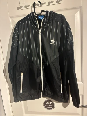 Svart vindjacka från Adidas - Svart vindjacka från Adidas köpt för 1200 kmr i adidas butiken. med vita tunna ränder och vit dragkedja. Jackan har huva, två fickor med dragkedja och Adidas logga på bröstet. Sportig och stilren design i lätt material, perfekt för streetwear.