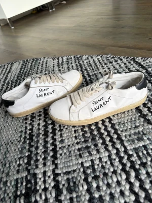 Saint Laurent vita låga sneakers - Vita sneakers från Saint Laurent med svart detalj på hälen och beige sula. Har dessvärre hål som syns på bilderna. Storlek 42. Skicka pm vid funderingar😊🙌